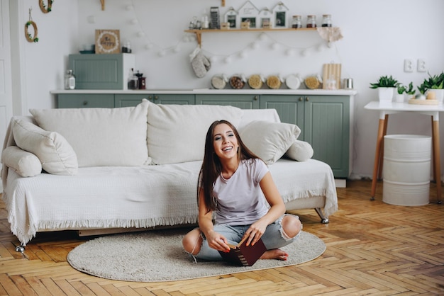 La fille s'assied sur le plancher près du sofa sur le fond de l'intérieur tient le livre dans ses mains