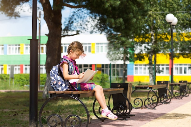 Fille avec un sac à dos assise sur un banc et lisant un livre près de l'école. Retour à l'école, horaire des cours, un journal avec les notes. Éducation, classes de primaire, 1er septembre