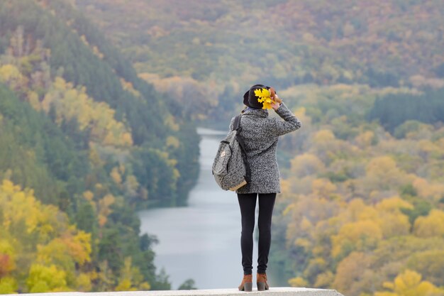 Fille avec un sac à dos et un chapeau debout sur une colline. Rivière et montagnes en contrebas. Vue arrière