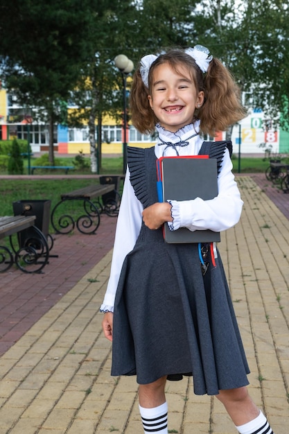 Fille avec sac à dos uniforme scolaire avec des arcs blancs et une pile de livres près de l'école Retour à l'école élève heureux manuels lourds Éducation classes d'école primaire 1er septembre