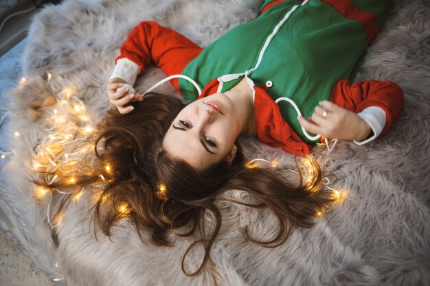 Une fille en salopette du Nouvel An avec une ambiance festive est allongée sur un lit autour de guirlandes lumineuses