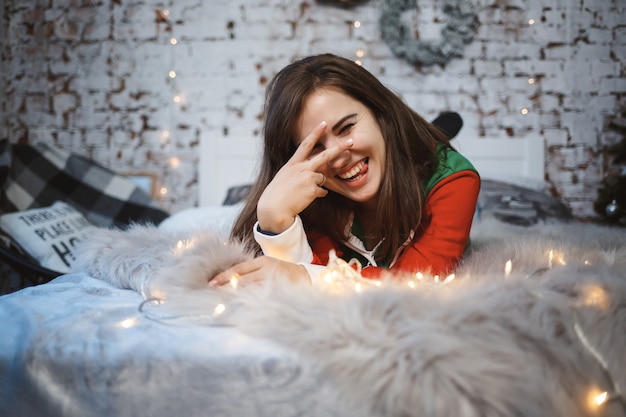 Une fille en salopette du Nouvel An avec une ambiance festive est allongée sur un lit autour de guirlandes lumineuses