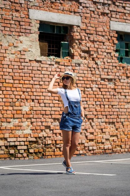 Fille en salopette en jean, un T-shirt blanc et un chapeau léger sur fond d'un bâtiment en brique sous le soleil éclatant