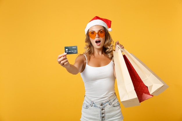 Fille de Santa choquée dans des lunettes de chapeau de Noël posant isolé sur fond jaune. Bonne année 2020 concept de vacances de célébration. Maquette de l'espace de copie. Tenez le sac après l'achat de la carte bancaire de crédit.