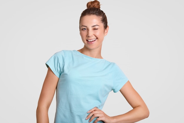 Une fille satisfaite aux yeux bleus clignote devant la caméra, garde une main sur la taille, a un chignon, vêtue d'un t-shirt décontracté bleu, pose contre le blanc, flirte avec l'homme. Hé, viens ici.