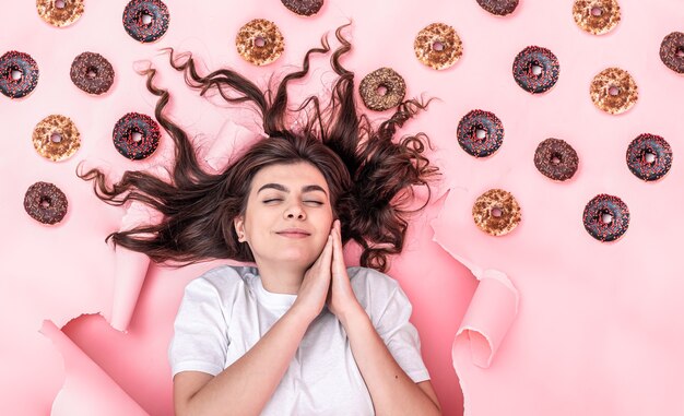 Une fille satisfaite aux yeux fermés sourit, allongée sur un fond de papier rose avec des beignets.