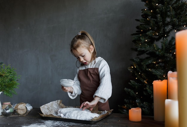 Une fille saupoudre du sucre en poudre sur un biscuit de Noël à une table de cuisine décorée de bougies