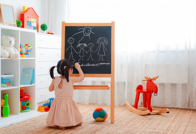 fille se tient dans la chambre des enfants avec un tableau noir et dessine une famille à la craie.