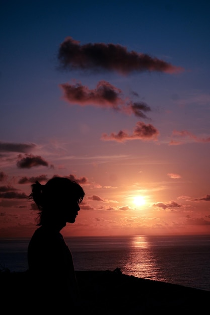 Une fille se tient devant un coucher de soleil avec le soleil se couchant derrière elle.