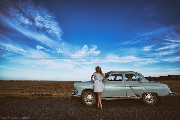 La fille se tient près d'une voiture rétro, du champ d'arrière-plan et d'un ciel bleu clair