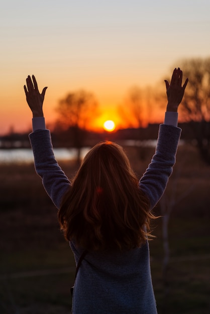 Fille se tient avec son dos pendant le coucher du soleil et lève les mains