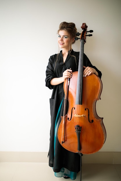 Une fille se tient avec un violoncelle contre un mur blanc un portrait en pied d'une femme violoncelliste