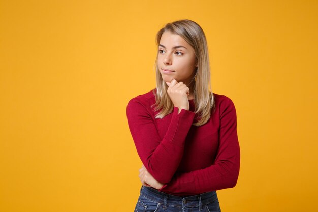 Fille séduisante jeune femme blonde dans des vêtements décontractés posant isolé sur fond orange jaune portrait en studio. Concept de style de vie des gens. Maquette de l'espace de copie. Mettez la main sur le menton, en regardant de côté.