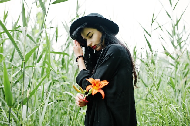 Fille sensuelle tout en lèvres rouges noires et chapeau Goth dramatique femme tient une fleur de lys orange sur un roseau commun