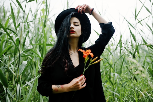 Fille sensuelle tout en noir, lèvres rouges et chapeau. Goth dramatique femme tenir fleur de lis orange sur le roseau commun.