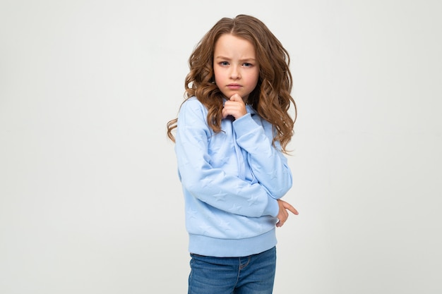 Fille sérieuse dans un chandail à capuchon bleu décontracté se dresse avec une expression honnête sur un mur blanc avec un espace vide