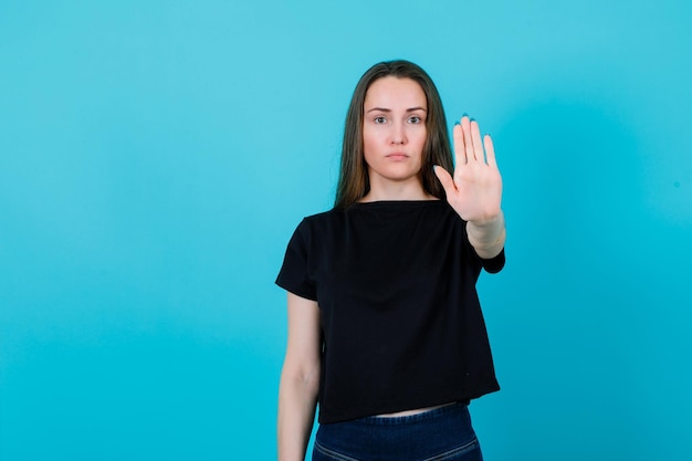 Une fille sérieuse montre un geste d'arrêt en étendant la main à la caméra sur fond bleu