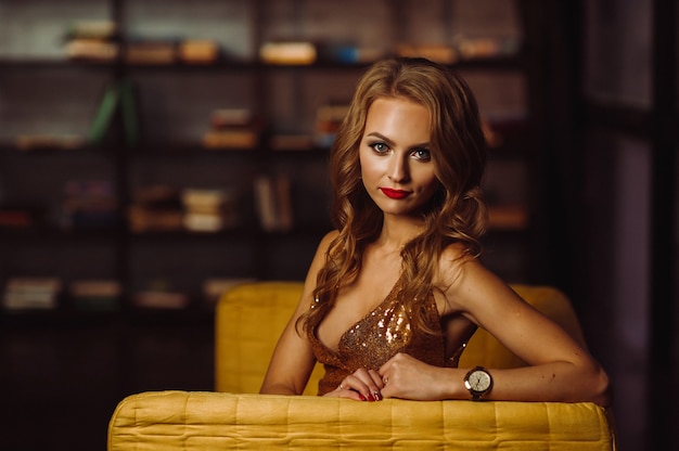 Fille sexy dans une robe dorée sur fond de livres dans la bibliothèque.Modèle aux cheveux longs et rouge à lèvres à l'intérieur.