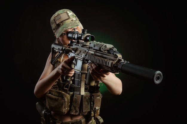 Une fille sexy en salopette militaire d'airsoft pose avec une arme à feu dans ses mains sur un fond sombre