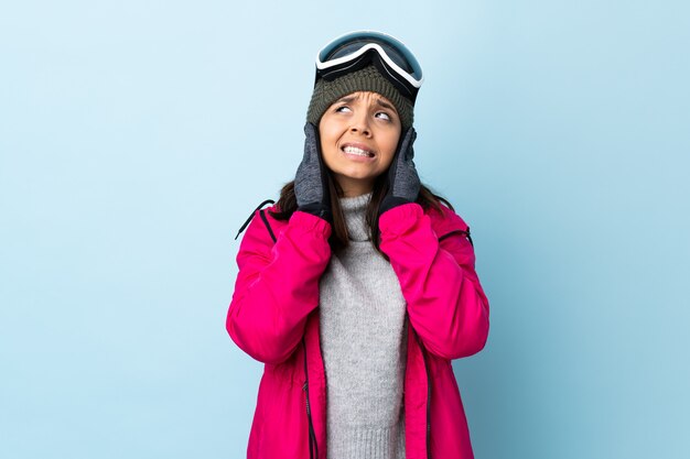 Fille de skieur de race mixte avec des lunettes de snowboard sur bleu isolé frustré et couvrant les oreilles.