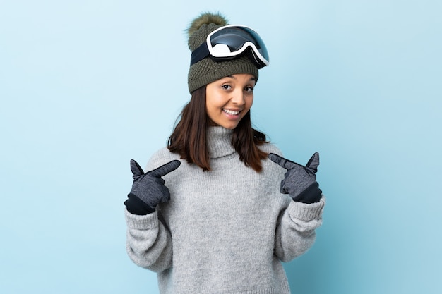 Fille de skieur de race mixte avec des lunettes de snowboard sur mur bleu isolé donnant un coup de pouce geste