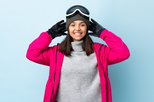 Fille de skieur de race mixte avec des lunettes de snowboard sur un mur bleu isolé en riant.