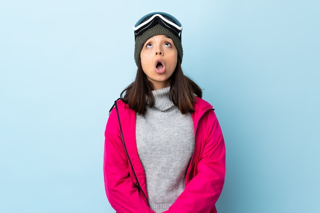 Fille de skieur de race mixte avec des lunettes de snowboard sur mur bleu en levant et avec une expression surprise