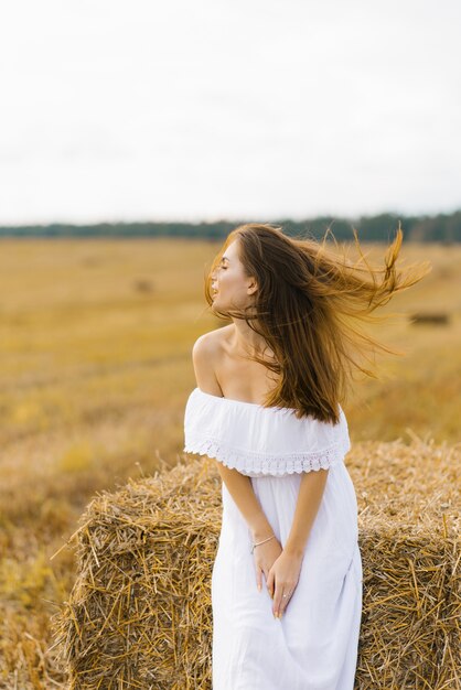 Fille slave aux longs cheveux blonds dans une robe blanche debout dans un champ avec des balles de paille, ses cheveux dans le vent