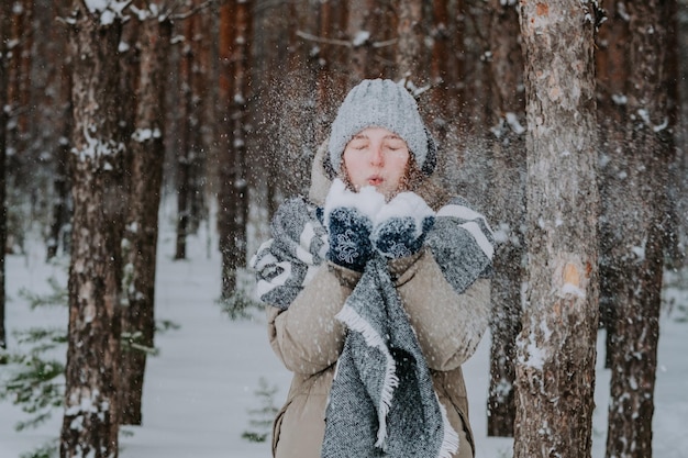 La fille souffle la neige de ses mains. Une fille au chapeau jette de la neige dans la forêt d'hiver. Bonnes vacances d'hiver. L'amour pour l'hiver. L'hiver dans le nord.