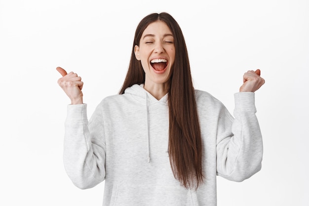 Une fille soulagée et heureuse crie d'excitation et de joie, serre la main les yeux fermés et rit, gagne, atteint l'objectif, célèbre le succès et gagne, debout sur un mur blanc