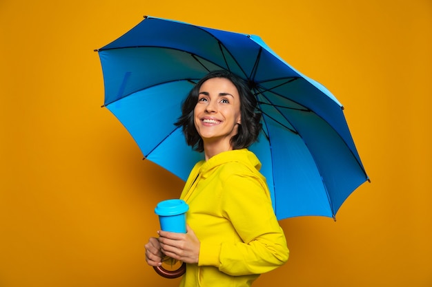 fille souriante dans une veste jaune sous un parapluie, tenant une tasse thermique bleue dans sa main gauche, levant les yeux avec espoir, profitant de la pluie.