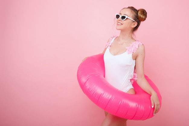 Fille souriante émotionnelle dans un maillot de bain blanc avec anneau en caoutchouc d'eau rose et lunettes sur un espace lumineux rose. humeur d'été