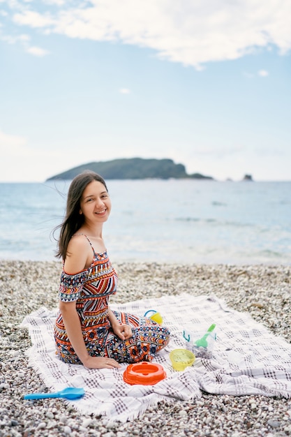 Une fille souriante est assise sur un couvre-lit sur une plage de galets avec des jouets pour enfants