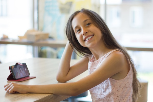 Une fille souriante est assise dans un café et communique dans un messager à l'aide d'un téléphone portable en regardant la caméra