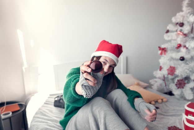 Une fille souriante est assise sur un lit sur fond d'arbre de Noël et donne un jouet