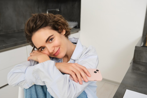 Fille souriante féminine assise sur une chaise de cuisine avec un smartphone rêvassant et semblant heureuse