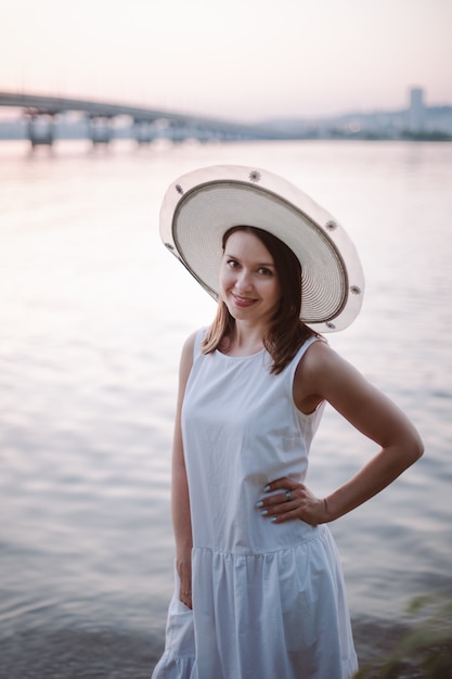 Une fille souriante heureuse dans une robe et un chapeau de paille blanc pose avec sa main sur sa taille sur une rivière être...