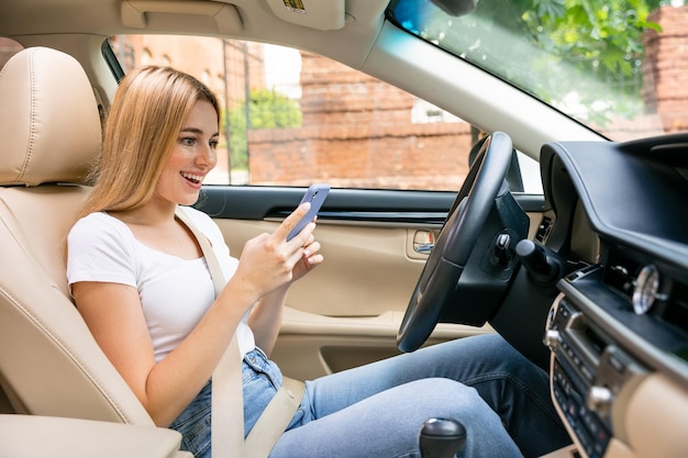 Une fille souriante lisant un message dans un téléphone portable est assise sur le siège du conducteur dans une voiture