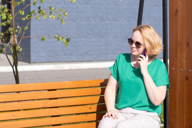 Une fille souriante à lunettes de soleil parlant au téléphone avec des amis assis sur un banc dans la rue