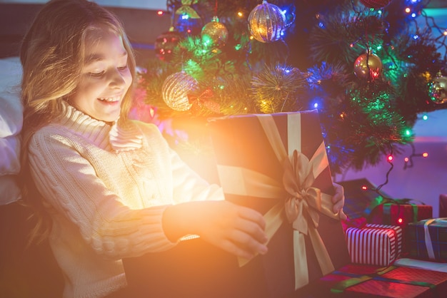 La fille souriante ouvre la boîte à lumière près de l'arbre de Noël. le soir la nuit