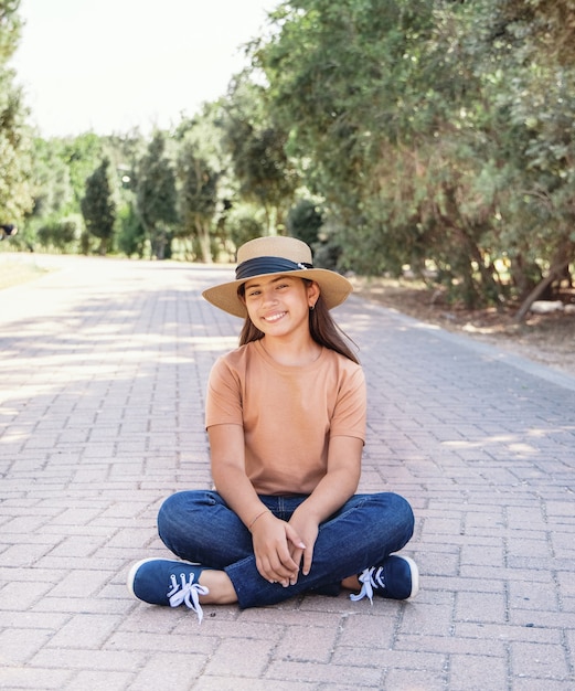 Une fille souriante portant un t-shirt, un jean et un chapeau de paille est assise en plein air dans le parc