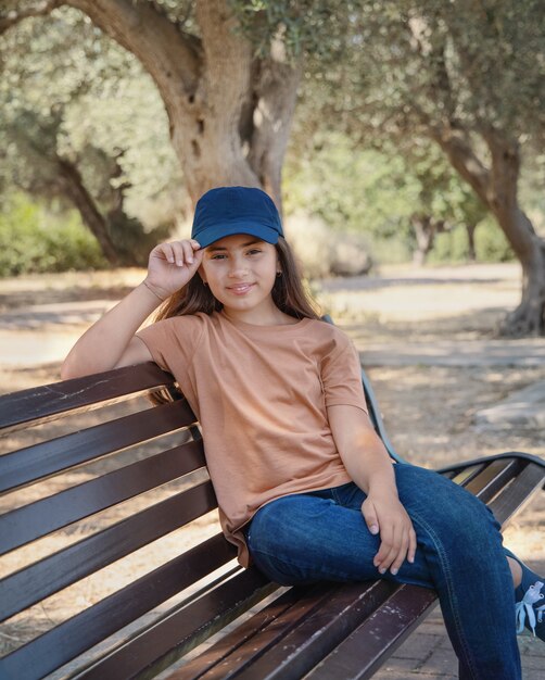 Fille souriante portant un t-shirt ; jeans et casquette de baseball est assis sur le banc dans le parc