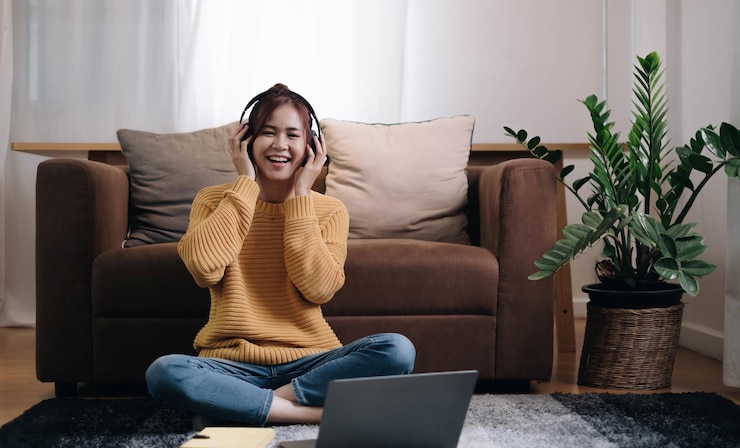 Photo fille souriante regardant la caméra à côté de l'ordinateur portable s'asseoir sur le sol devant le canapé et mettre des écouteurs pour écouter de la musique à la maison