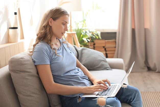 Une fille souriante s'assoit sur un canapé en regardant un webinaire sur un ordinateur portable. Heureuse jeune femme étudie sur un cours à distance en ligne.
