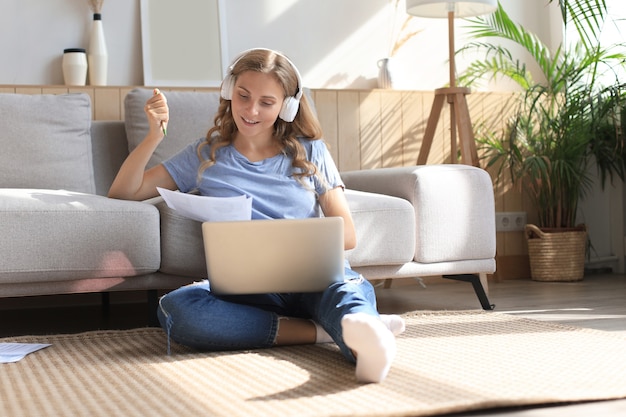 Une fille souriante s'assoit près du canapé en regardant un webinaire sur un ordinateur portable. Heureuse jeune femme étudie sur un cours à distance en ligne.