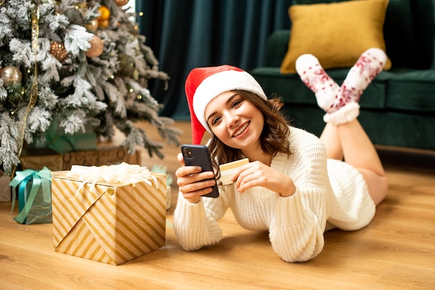 Fille souriante avec smartphone et carte de crédit près de l'arbre de Noël. Achats en ligne le nouvel an. Elle achète des cadeaux