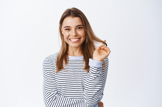 Fille souriante et vous regardant avec un visage pensif, debout sur un mur blanc