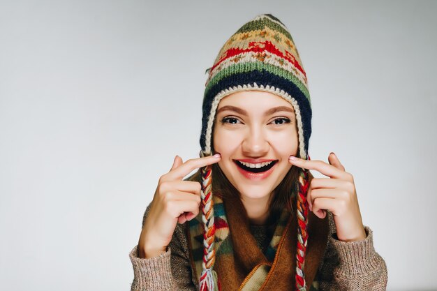 La fille sourit et montre sur ses joues, portant un chapeau et une écharpe avec un ornement scandinave