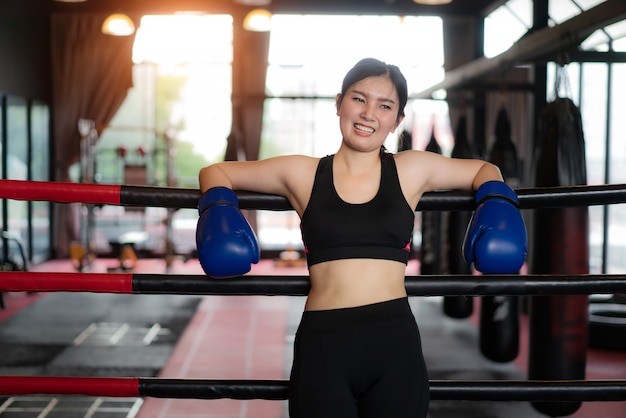 Fille de sport boxeur asiatique fatigué s'appuya sur des cordes rouges noires sur le ring de boxe et se repose après un entraînement dur dans une salle de sport loft noir. Ajustement sportif pour un mode de vie sain Modèle asiatique du concept de gym de boxe.