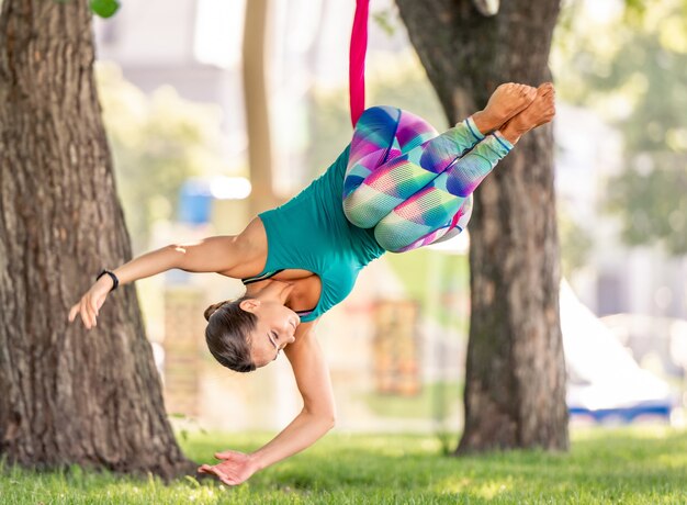 Fille sportive pratiquant le yoga à la mouche dans un hamac dans la nature et gardant l'équilibre dans les airs avec la tête en bas. Jeune femme faisant de la gymnastique aérodynamique qui s'étend à l'extérieur